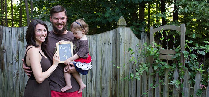 pregnant mom with dad and their young daughter holding chalkboard sign that says party of 4 in front of a garden gate