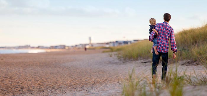 Image of young dad holding his toddler as they walk along a sandy beach