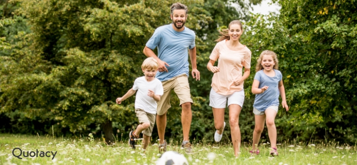 Family of four running through a field with pine trees in the background chasing a soccer ball