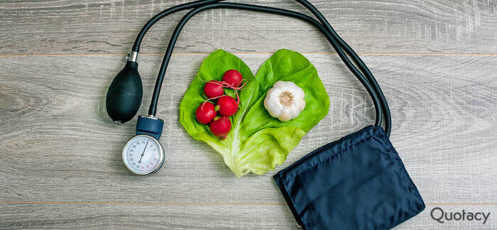 A blood pressure cuff next to a heart shaped piece of lettuce with radishes on one side and a bulb of garlic on the other all of which sits on a gray wooden table