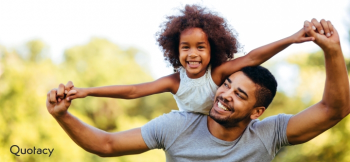 Father holding daughter on his shoulders with trees in the background.