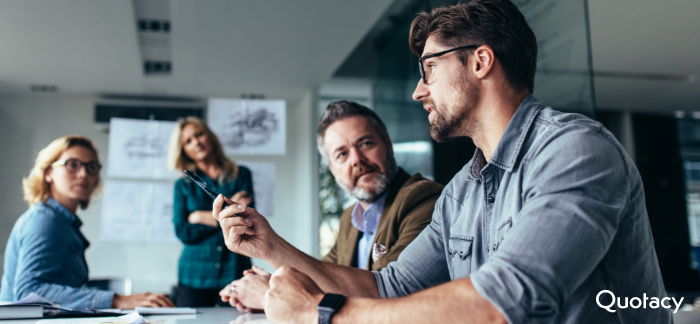 Image of a business man in a blue button up talking to another man and two women in a conference room.