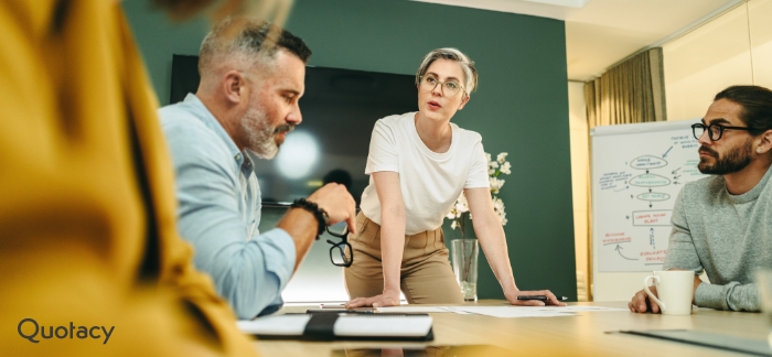 An image of a woman standing at the head of a table in a conference room with three other people sitting at the table looking at her intently. Behind the woman at the head of the table is a whiteboard with business drawings on it.