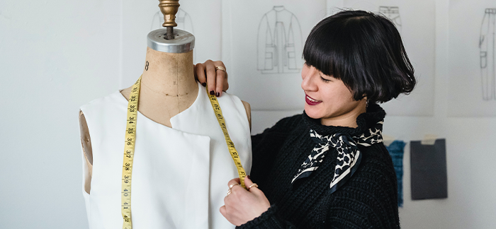 Woman of Asian decent holding a fabric tape measurer up to a white dress on a dummy